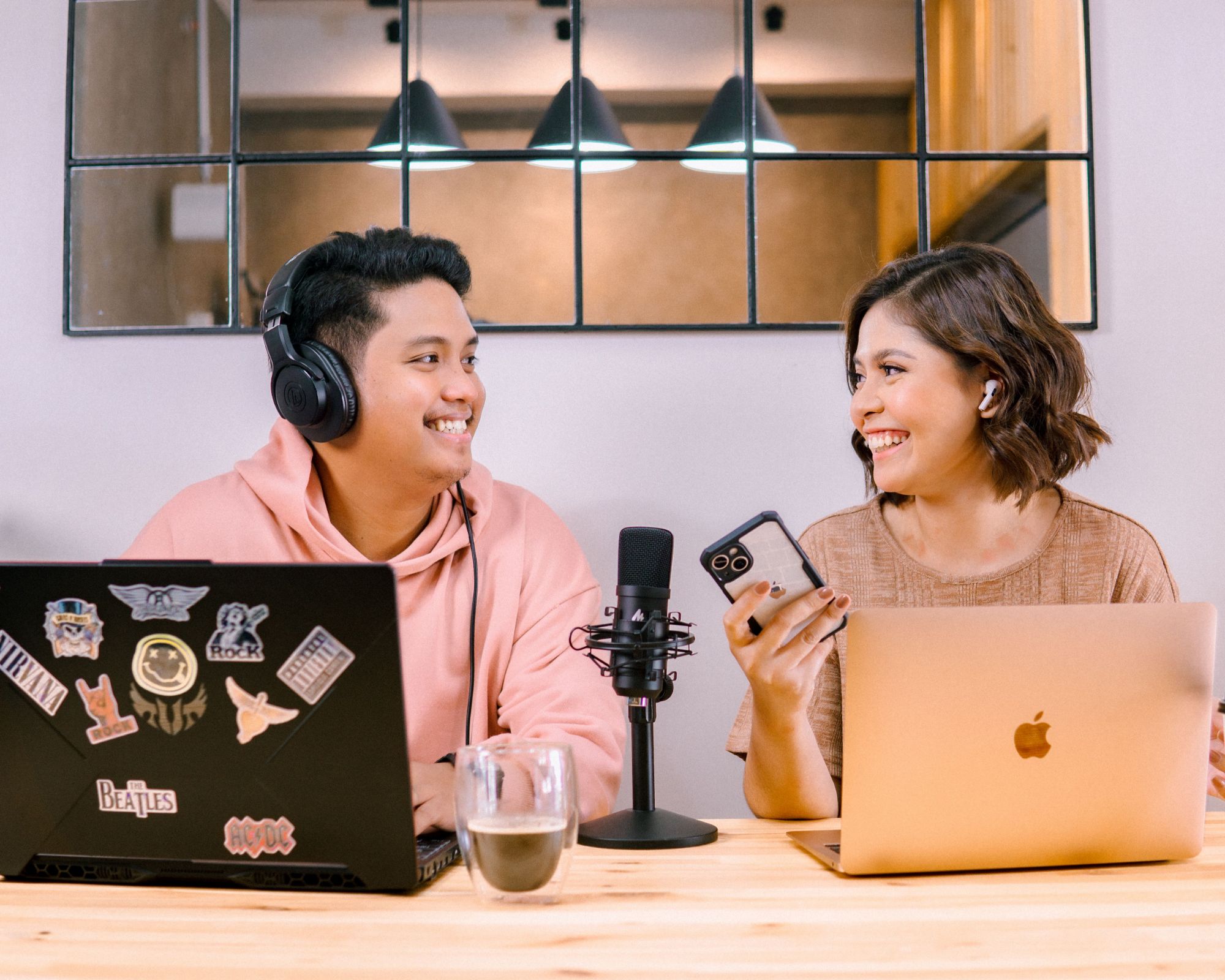 Two radio hosts smiling at each other across a studio desk with laptops and a microphone